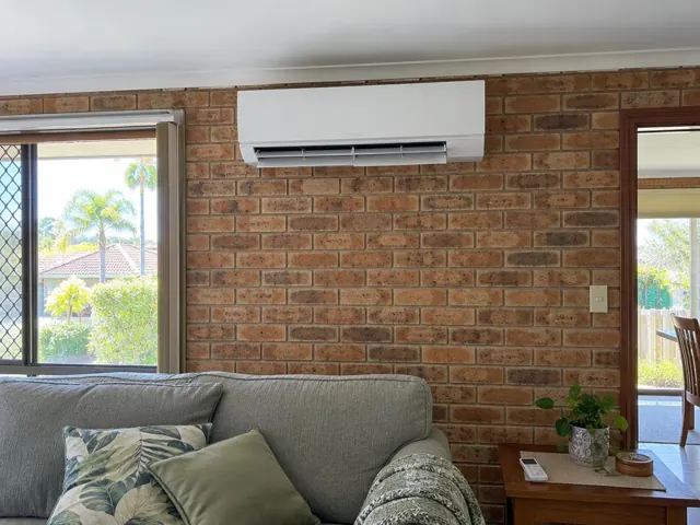 Wall-mounted split system running on a brick wall in a living room with a sofa and windows in Burleigh Waters