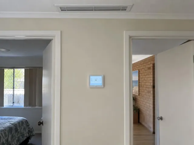 Ceiling ducted air vent and wall controller in a beige hallway between doorways in Robina