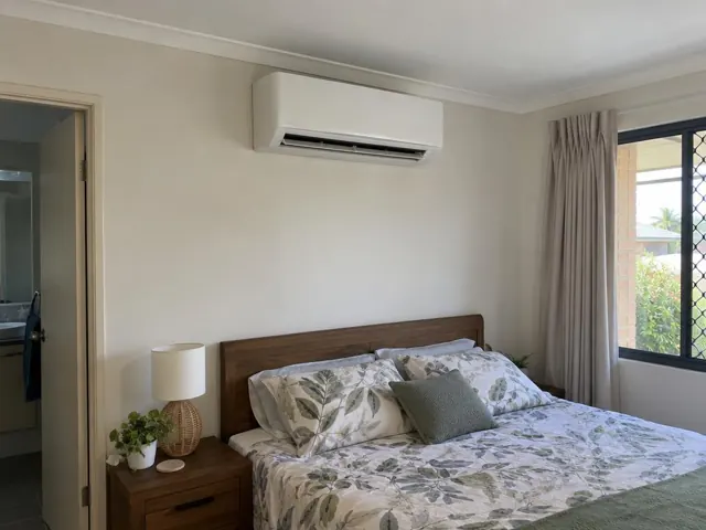 Wall-mounted split system above a bed in a bedroom with a window and bedside table in Varsity Lakes